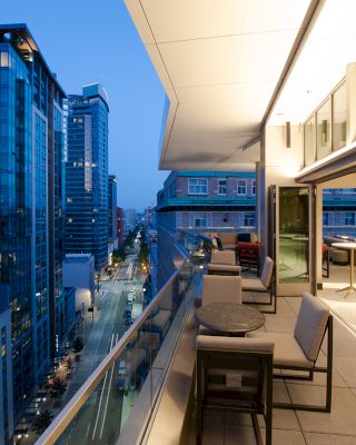 A modern balcony with seating and tables overlooks a cityscape with tall buildings and lit streets at dusk or early evening.