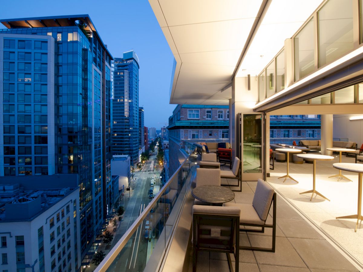 A modern outdoor balcony view of an urban skyline at dusk, featuring outdoor seating and tables against a backdrop of tall buildings.