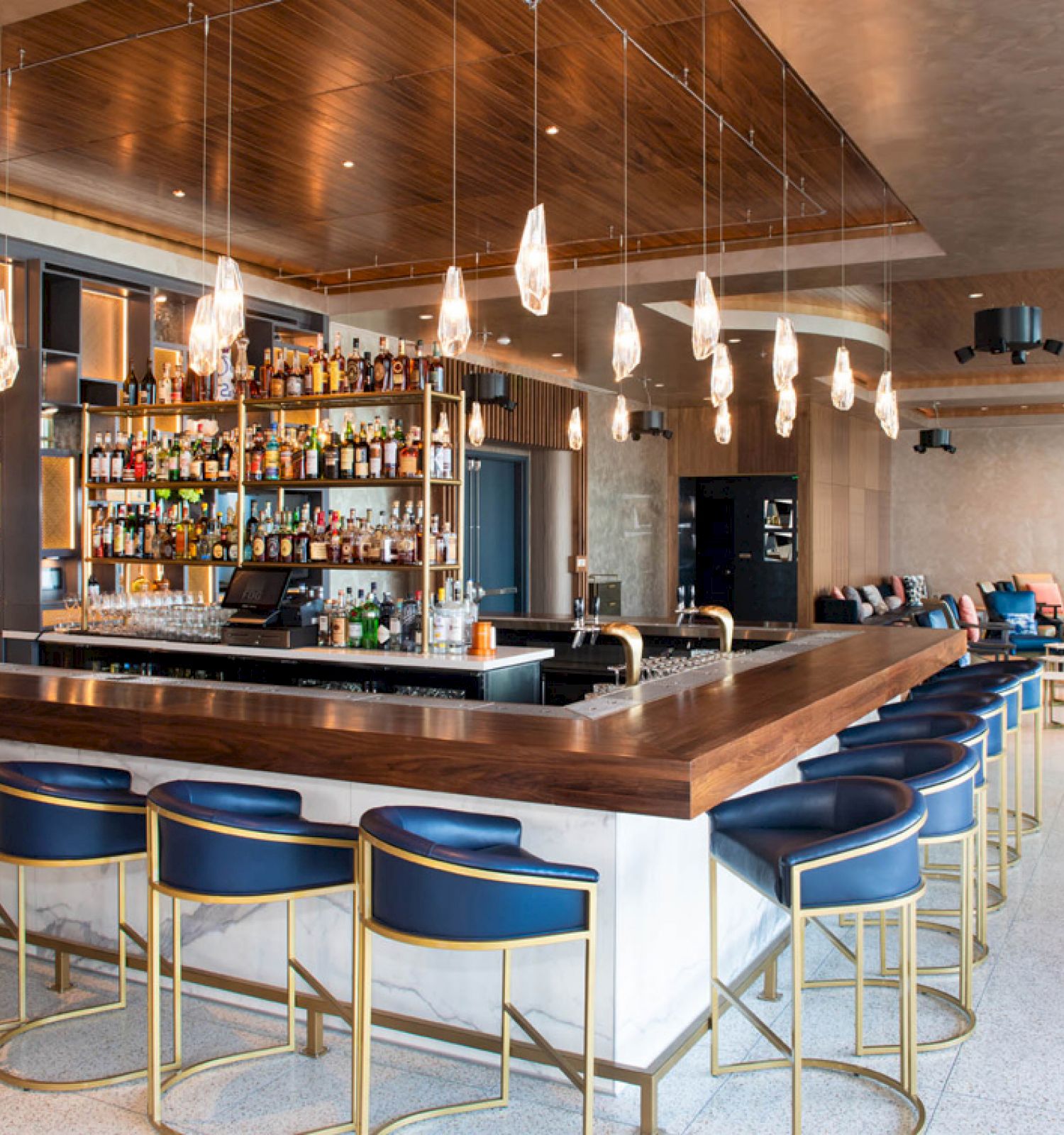 A modern bar with a stocked liquor shelf, hanging lights, and blue cushioned bar stools. There is additional seating in the lounge area.