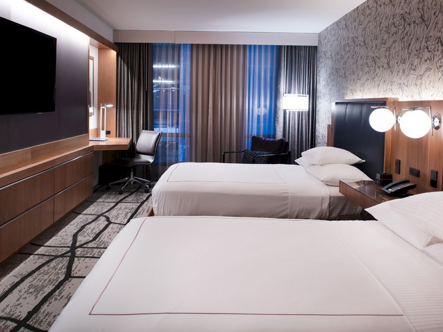 The image shows a modern hotel room with two neatly made beds, a desk with a chair, a flat-screen TV, and contemporary lighting.