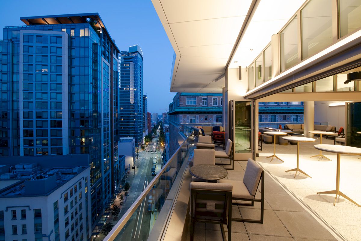 A modern balcony overlooks a street lined with tall buildings at dusk. The balcony has large glass doors and is furnished with tables and chairs.
