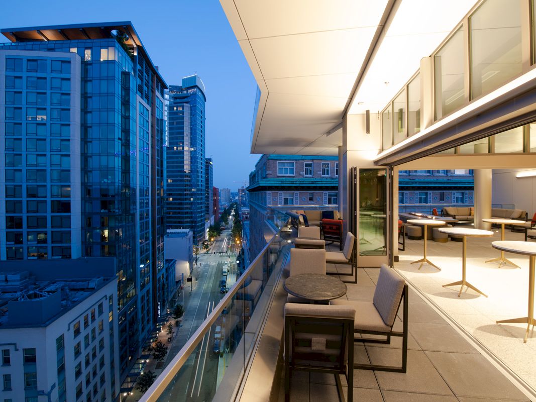 A modern balcony overlooks a street lined with tall buildings at dusk. The balcony has large glass doors and is furnished with tables and chairs.