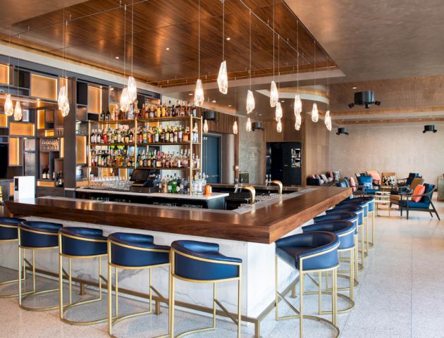 A modern bar with blue stools around a wooden counter, adorned with hanging lights. The back wall has shelves filled with bottles and glasses.