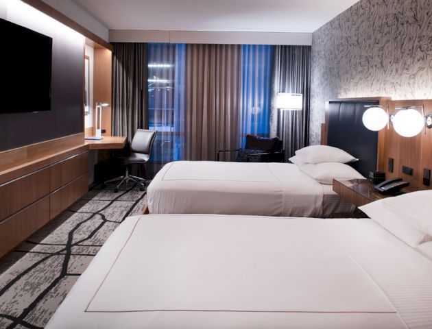 The image shows a modern hotel room with two neatly made twin beds, a TV, a desk with a chair, and wall-mounted lamps.