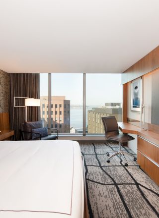 A modern hotel room with a bed, TV, desk, chair, large window showing a city view, and stylish decor.