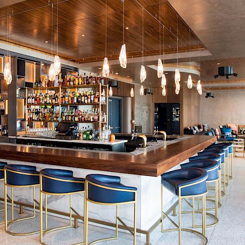 A modern bar with a wooden countertop, blue bar stools, hanging lights, and shelves stocked with bottles. Seating area visible in the background.