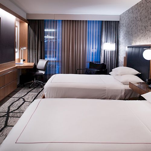 A modern hotel room with two neatly made beds, a desk, a TV, and a large window with curtains, featuring contemporary decor and lighting.