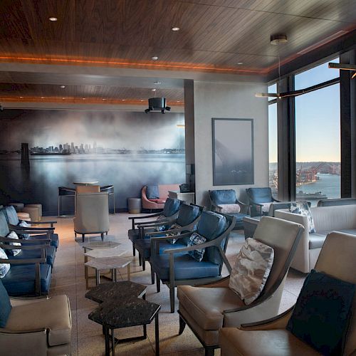 A modern lounge with leather chairs, tables, large windows offering a scenic view, and a minimalistic design with a cityscape wall mural, ending the sentence.