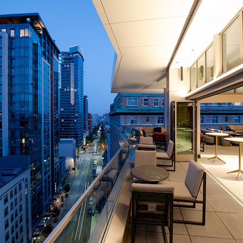A modern cityscape at dusk with tall buildings, viewed from the balcony of a stylish, open-air restaurant or lounge with tables and chairs.