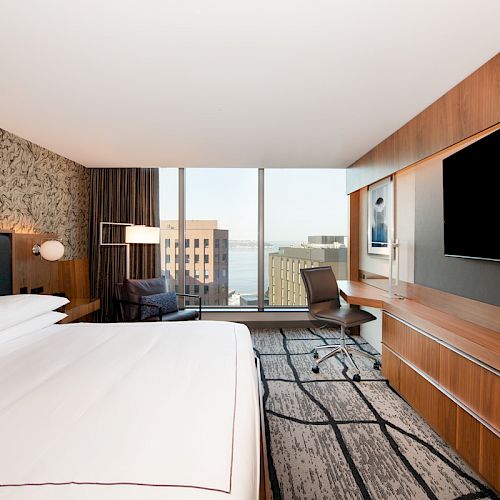 This image shows a modern hotel room with a king-sized bed, a large flat-screen TV, a desk, a chair, and a window with a cityscape view.