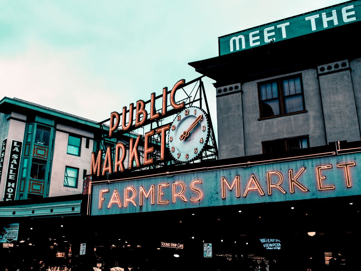 The image shows a neon sign reading "Public Market" and "Farmers Market" with a clock in between, located on the facade of a historic building.