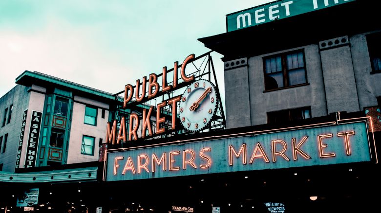 The image shows a neon sign reading "Public Market" and "Farmers Market" with a clock in between, located on the facade of a historic building.