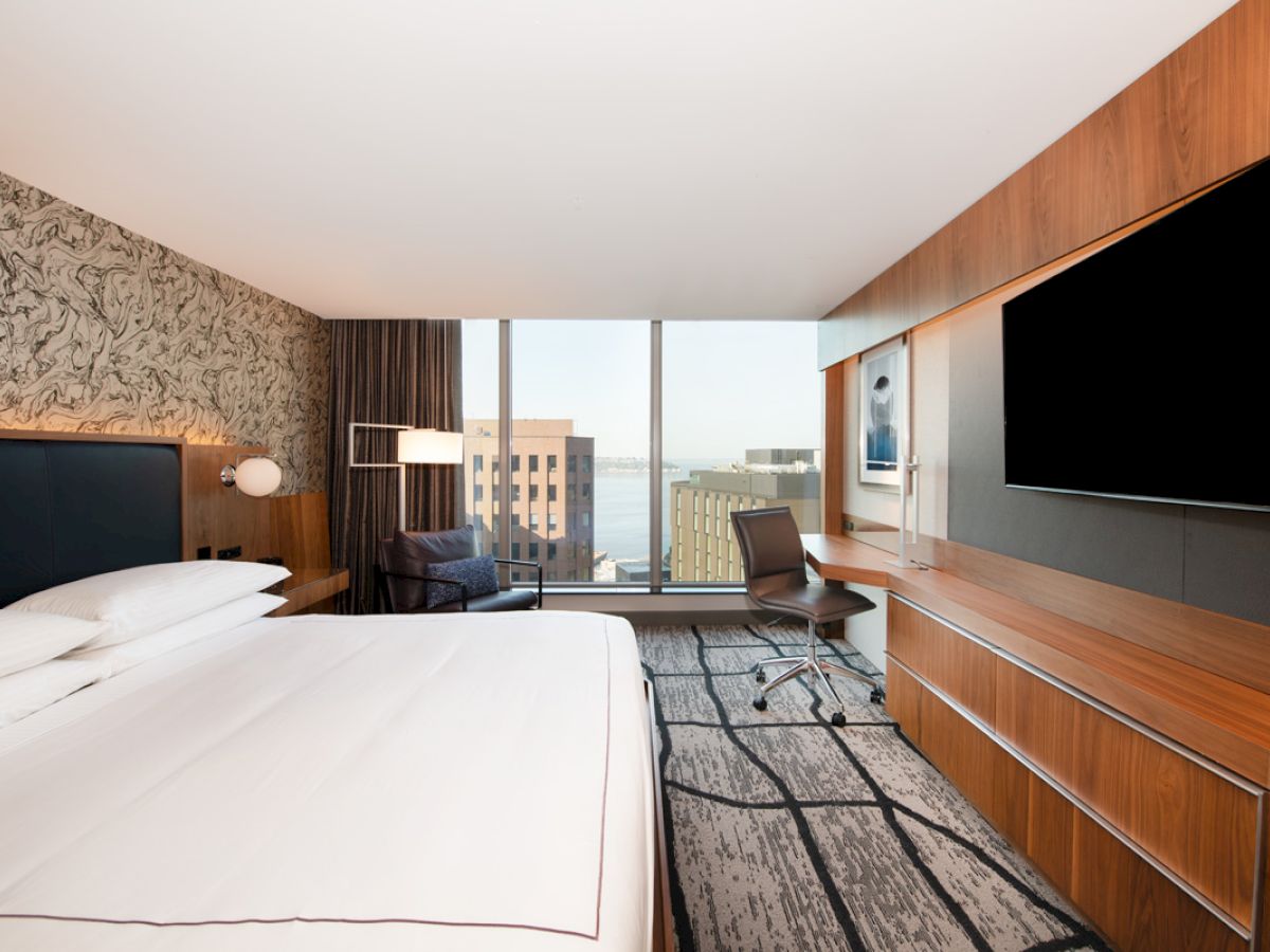 A modern hotel room featuring a large bed, wall-mounted TV, desk with a chair, large window with a city view, and contemporary decor.