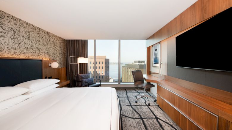 A modern hotel room featuring a large bed, wall-mounted TV, desk with a chair, large window with a city view, and contemporary decor.