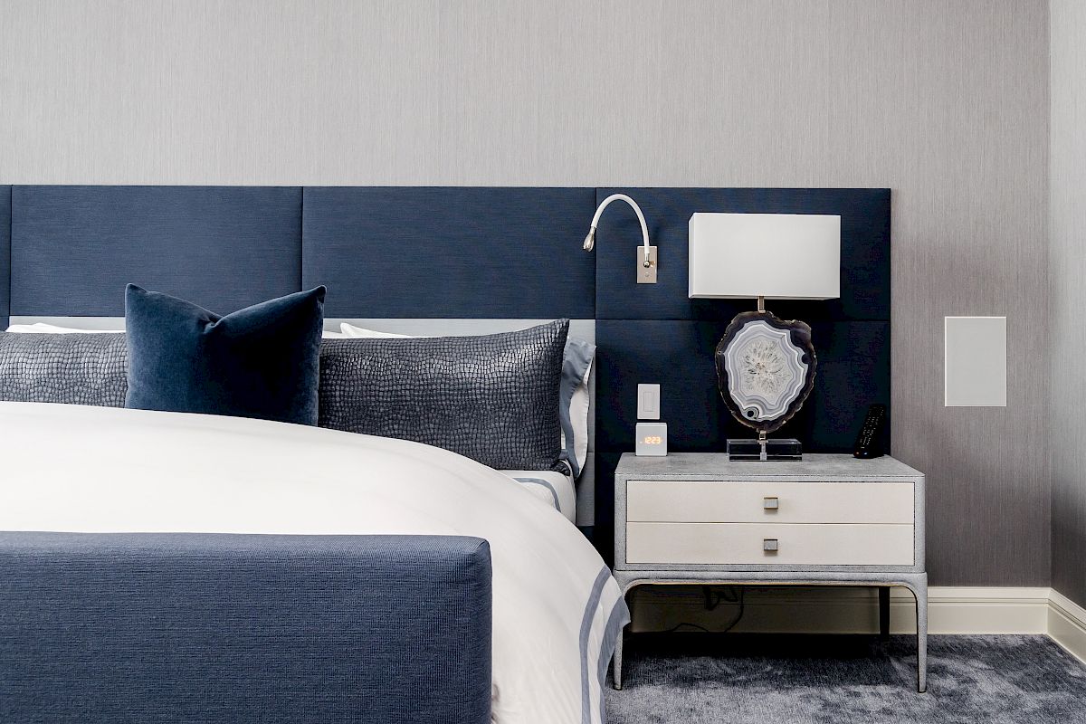 A modern bedroom with a blue and white color scheme features a bed, nightstand, lamp, and wall-mounted reading light.