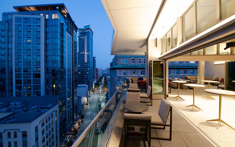 A modern outdoor balcony with seating and tables overlooks a city street lined with tall buildings at dusk.