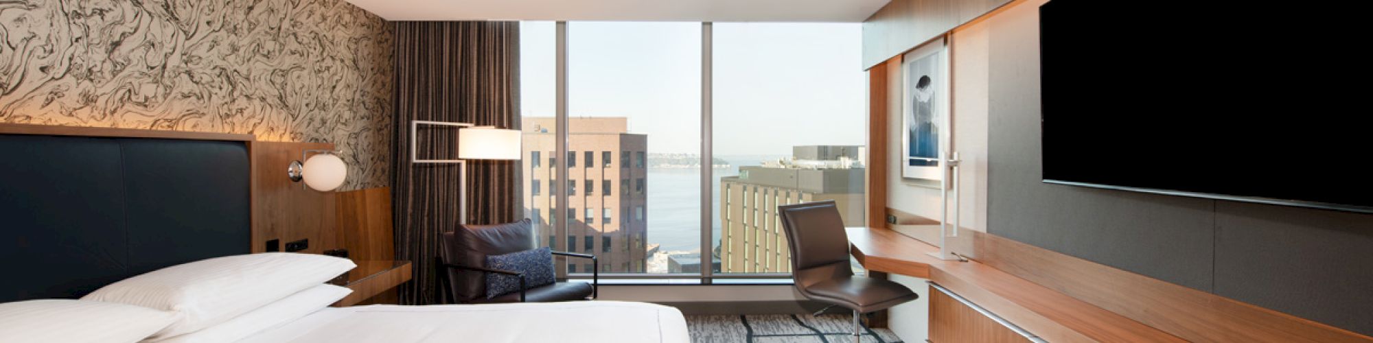 A modern hotel room with a large bed, artistic wallpaper, a desk with a chair, and a large TV. Floor-to-ceiling windows provide a city view.