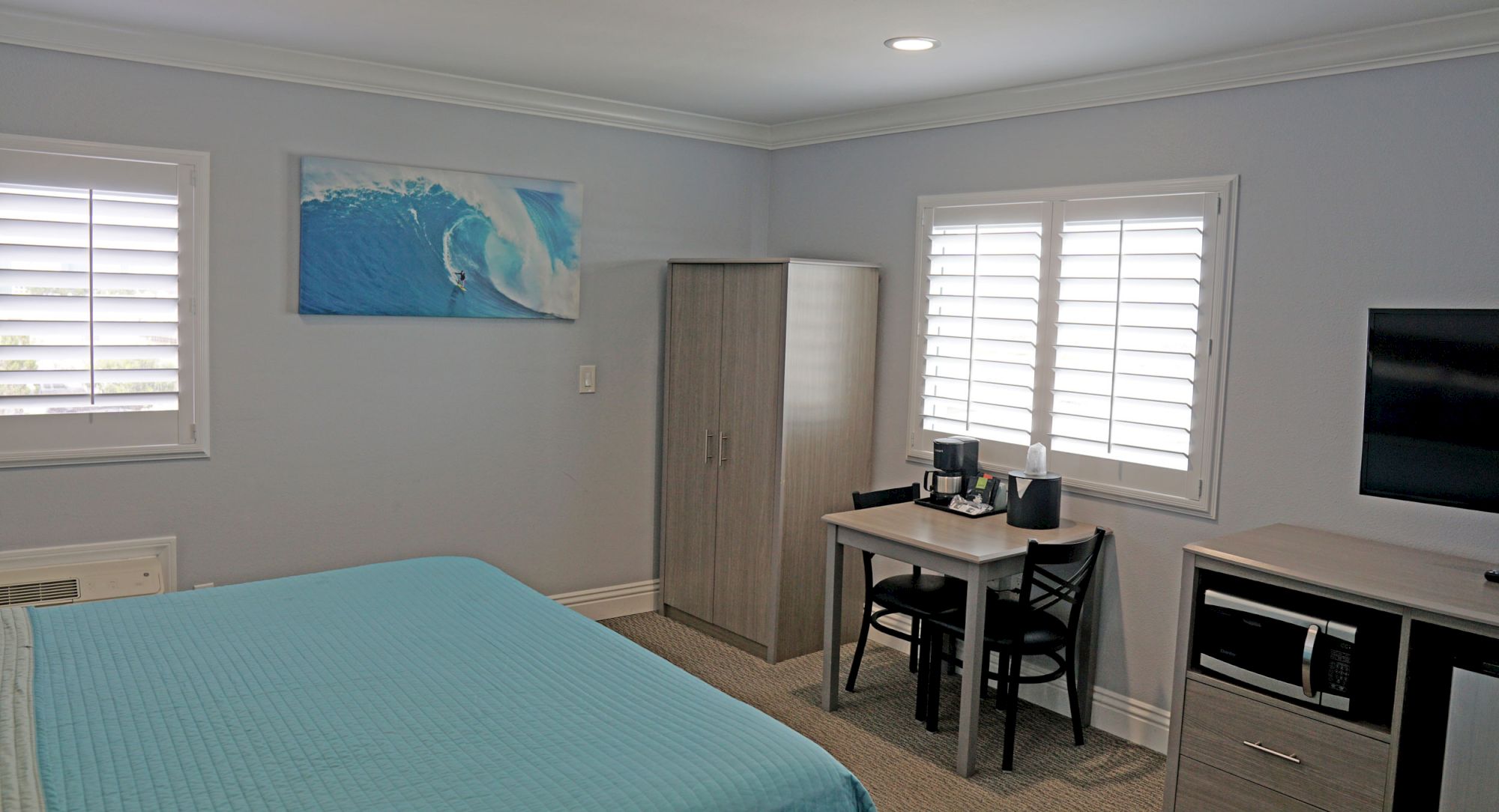 The image shows a bedroom with a bed, wardrobe, table, chairs, wall art of a wave, two windows with blinds, a TV, and a mini-fridge on a stand.