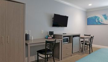 The image shows a hotel room with a TV on the wall, a small desk, a mini-fridge, a microwave, two chairs, and a picture of a wave on the wall.