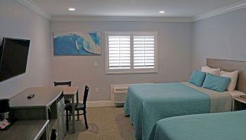 A hotel room with two beds, a TV, a small desk with chairs, a coffee maker, and wall art of a wave. The room is well-lit and has a window with shutters.