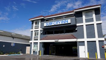 The image shows a building labeled "Surf City Inn" with a dark gray exterior and white trim, under a blue sky with clouds in the background.