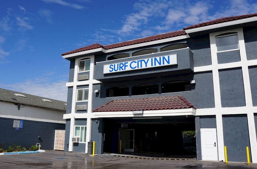 The image shows a building labeled "Surf City Inn" with a dark gray exterior and white trim, under a blue sky with clouds in the background.