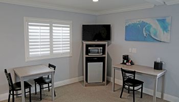 A room with two tables and chairs, a TV, mini-fridge, microwave, coffee maker, window with blinds, and a wall-mounted wave painting.