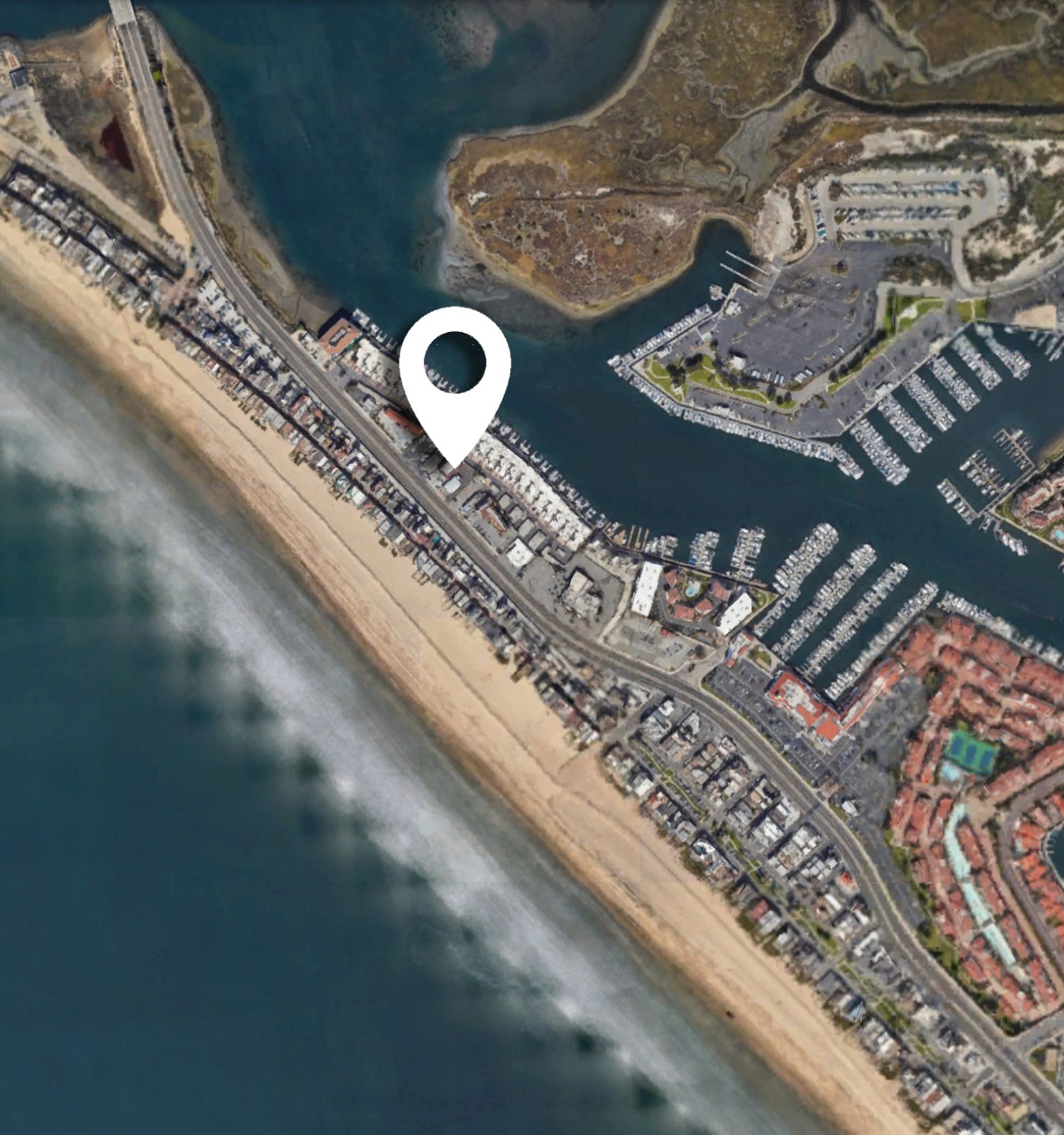 An aerial view of a coastal area featuring a beach, marina, and residential developments with a location pin marker placed on the image.