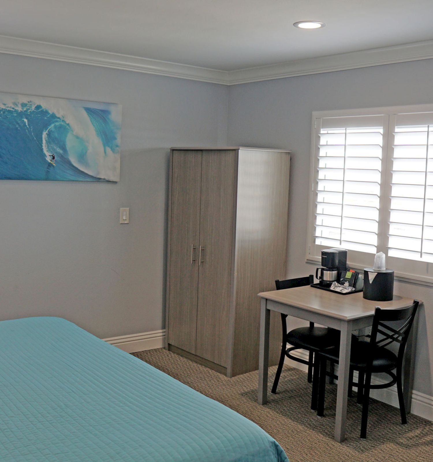 A modern bedroom with a bed, wardrobe, desk, chairs, TV, and a seascape painting. The room is bright with two shuttered windows and neutral decor.