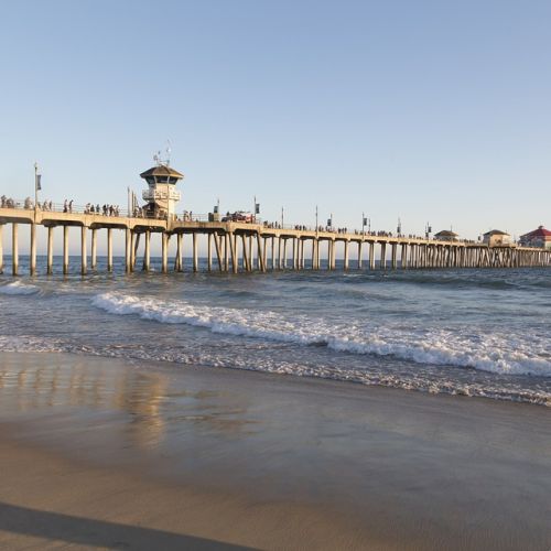 A pier stretches into the ocean as two people stand on the beach, facing the camera with waves gently touching the shore in the evening light.