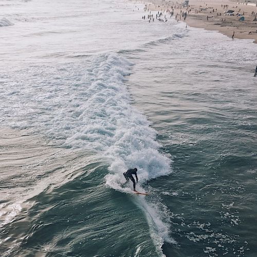 A lone surfer rides a wave towards a beach crowded with people, while the ocean stretches further into the horizon under a clear sky.