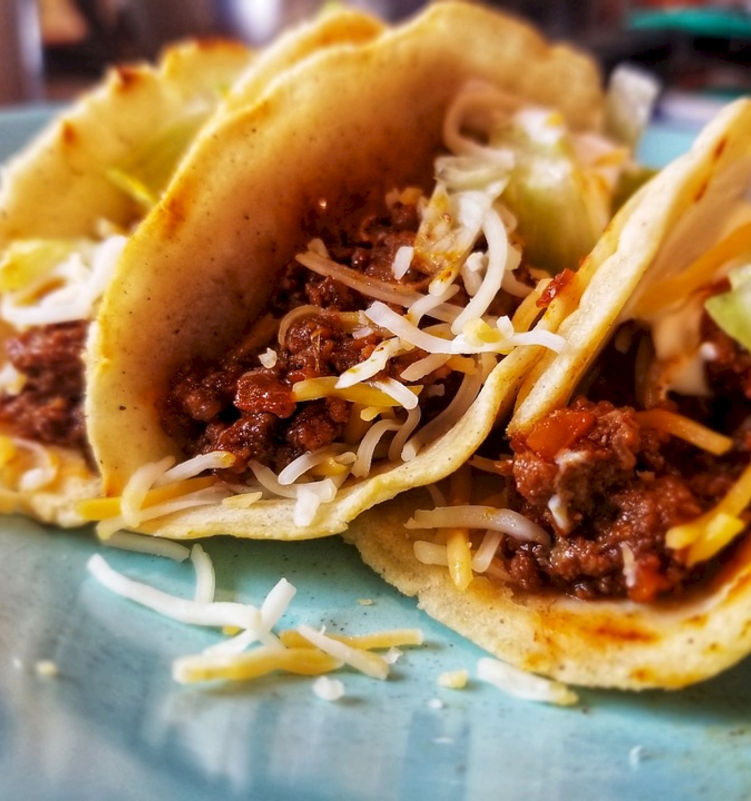 The image shows three tacos filled with meat, shredded cheese, and lettuce on a blue plate.