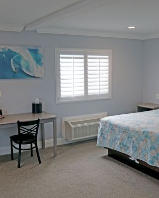 A tidy bedroom with a double bed, a desk with a chair, a TV, a microwave, a mini fridge, and a beach-themed painting on the wall.