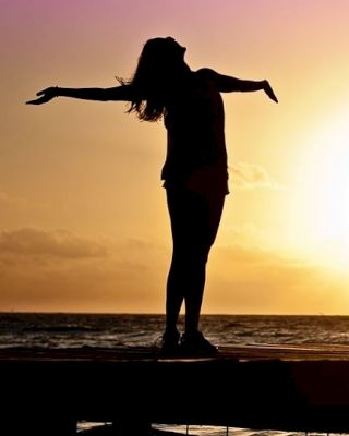 A silhouetted person stands with arms outstretched by the sea, against a vibrant sunset backdrop.