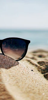 A pair of black sunglasses resting on a sandy beach with the ocean in the background, suggesting a sunny day at the seaside.